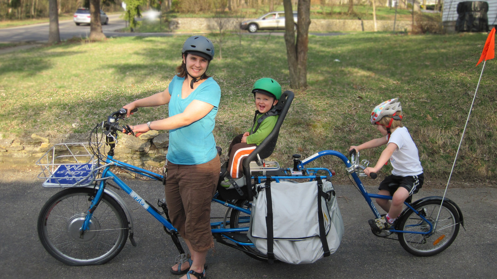 Mom stands next to electric cargo bike with two kids on board. One in child seat and one on attached trailerbike.