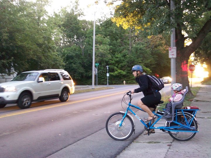 Woman in professional clothes turns into traffic to bike to work on a cargo bike. Baby is on the back. 