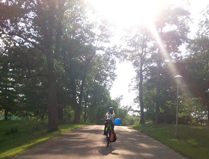 Mother rides with child through a park with sunny park. A blue balloon blows in the breeze from the bike. 