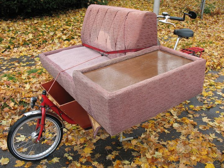 A small couch strapped across a cargo bike. Ground is covered by orange leaves. 
