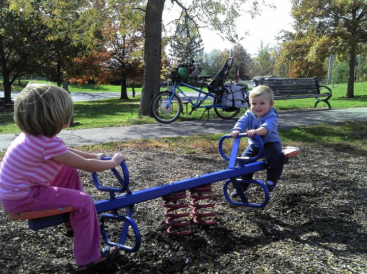 Two kids play on a teeter totter with a cargo bike in the background