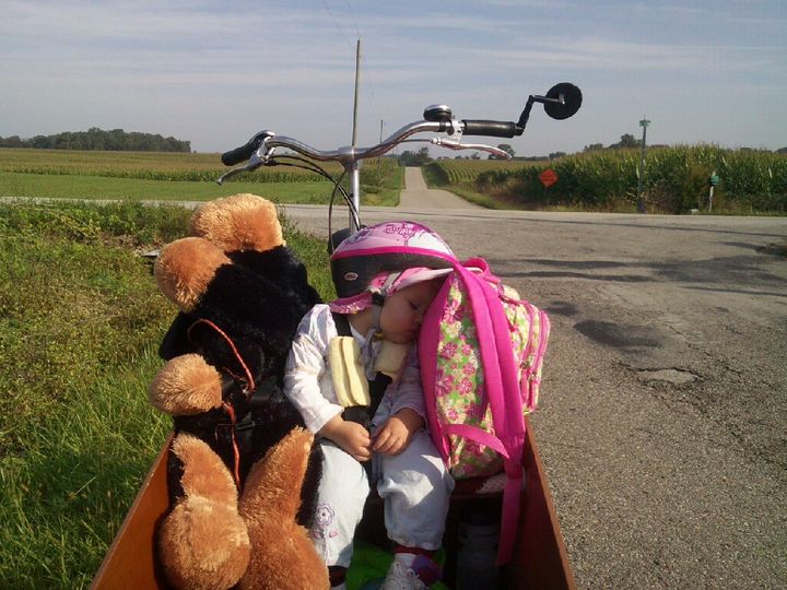 Child sleeps on a cargo bike in the countryside. Head rests on backpack. A child-sized stuffed animal rests next to her. 