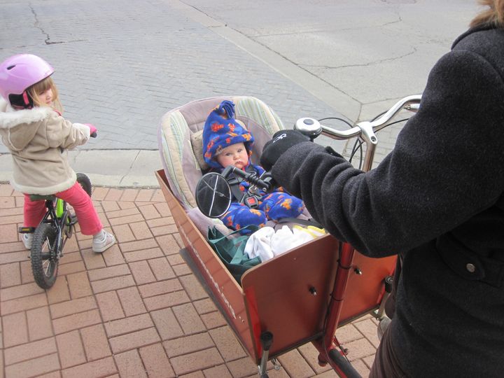 3 year old girl waits to cross a straight on a bike with her family. Baby brother is cozy in a bakfiets. 